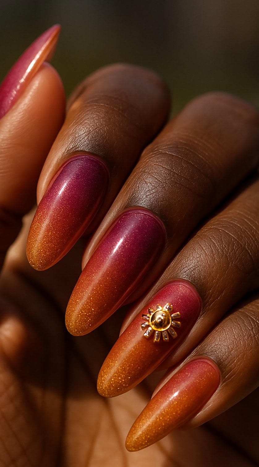 Long almond nails with a red-to-gold ombré shimmer and a golden sun charm on one nail.