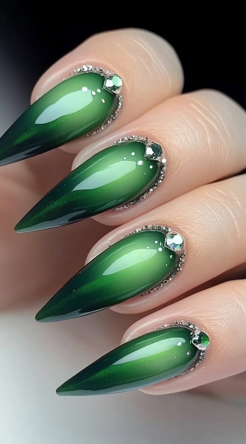 A hand with long stiletto-shaped nails featuring a deep green aura effect with rhinestones and metallic beads adorning the cuticle area.