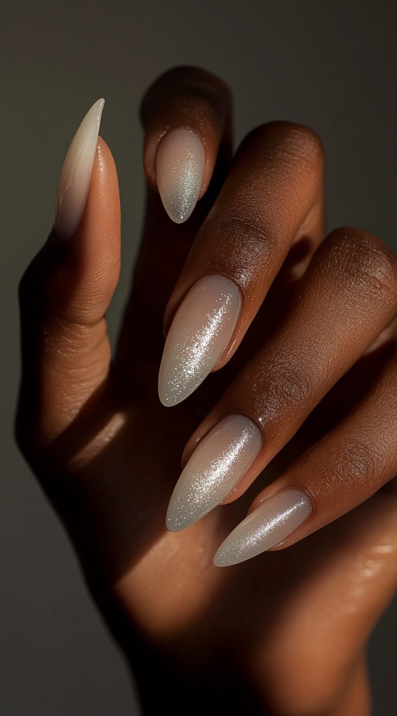 A close-up of a dark-skinned hand with almond-shaped nails painted in a sheer pinkish-nude base, topped with subtle fine glitter.