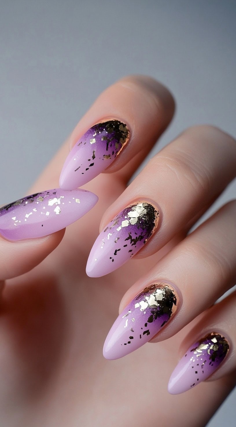 Almond nails painted in a lilac-to-violet ombre, adorned with black accents and metallic gold foil around the cuticles.