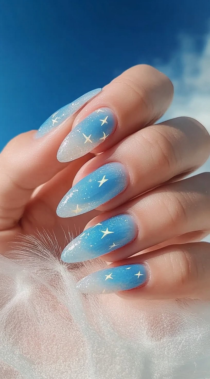 Almond-shaped nails with a light blue to white ombre effect and golden star accents.