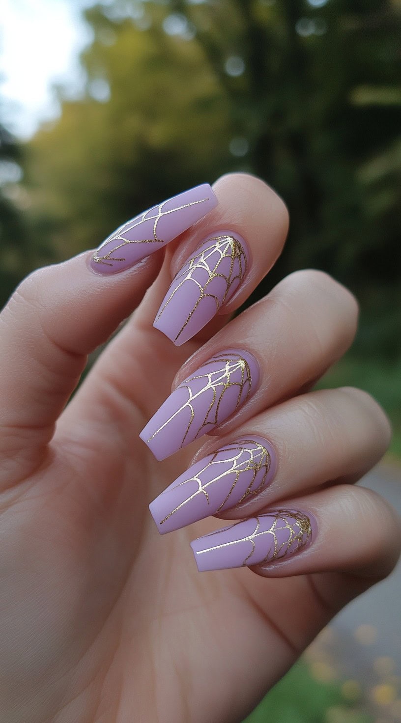 Coffin-shaped nails painted in soft lilac with intricate gold spiderweb line art across the surface.