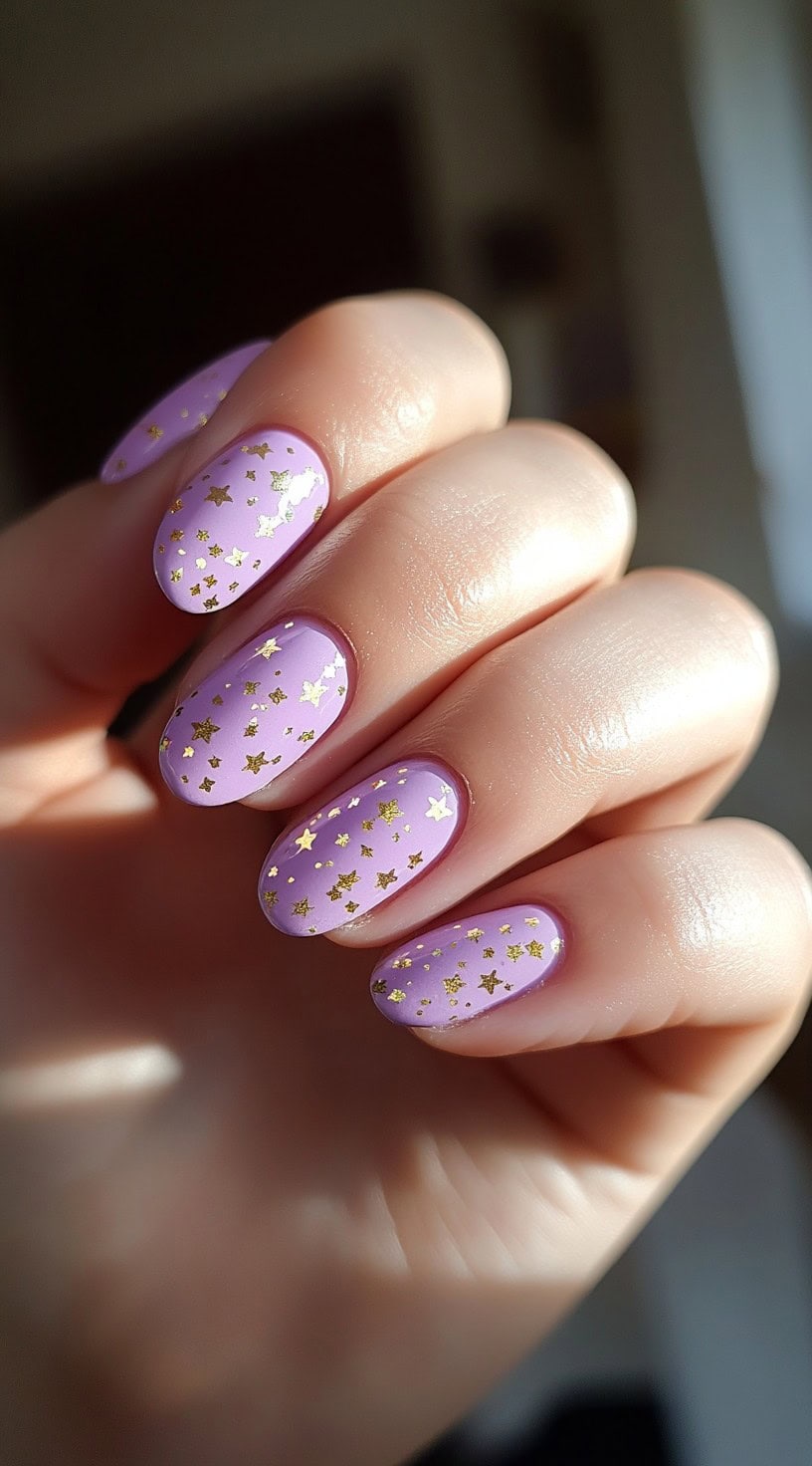 Short oval nails painted in soft lilac and covered in small gold star confetti scattered across each nail.