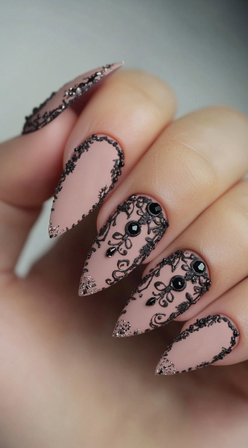 A hand with matte nude almond-shaped nails detailed with black lace patterns and small black rhinestones.