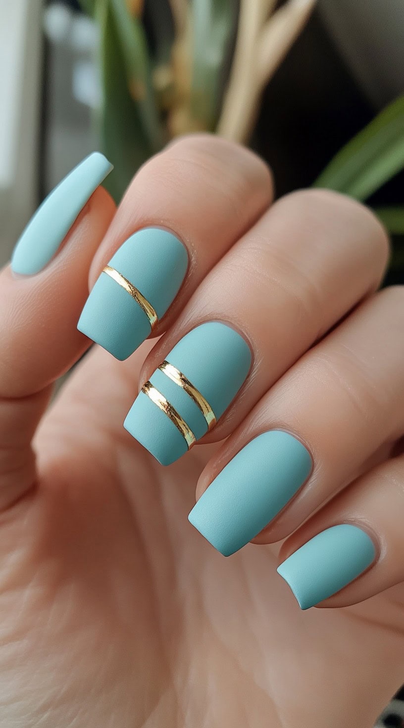 Matte light blue square nails with two horizontal gold stripes on the ring finger and middle finger.