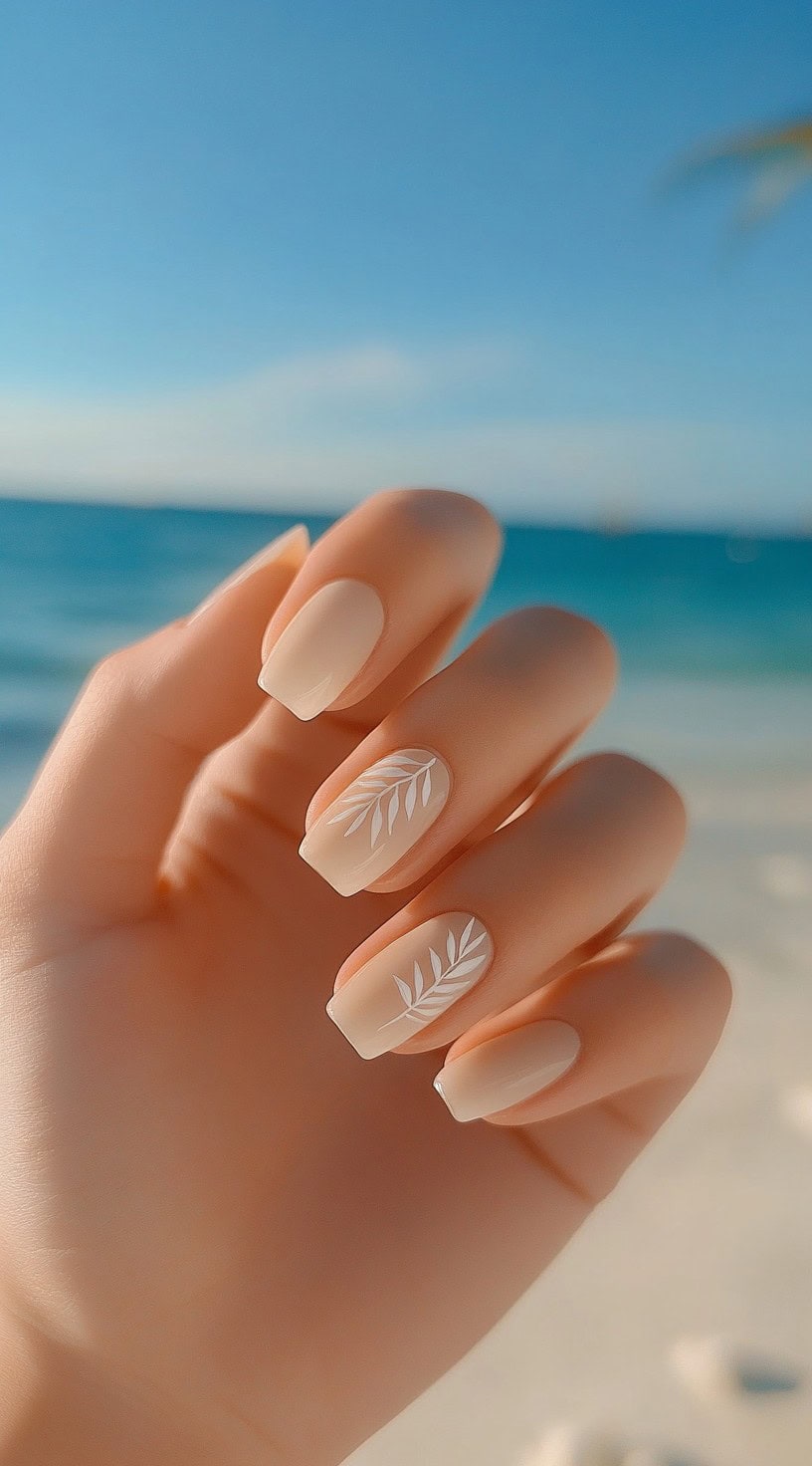 Square-shaped matte nude nails with a delicate white leaf design on two accent nails, framed by a soft, beachy background.