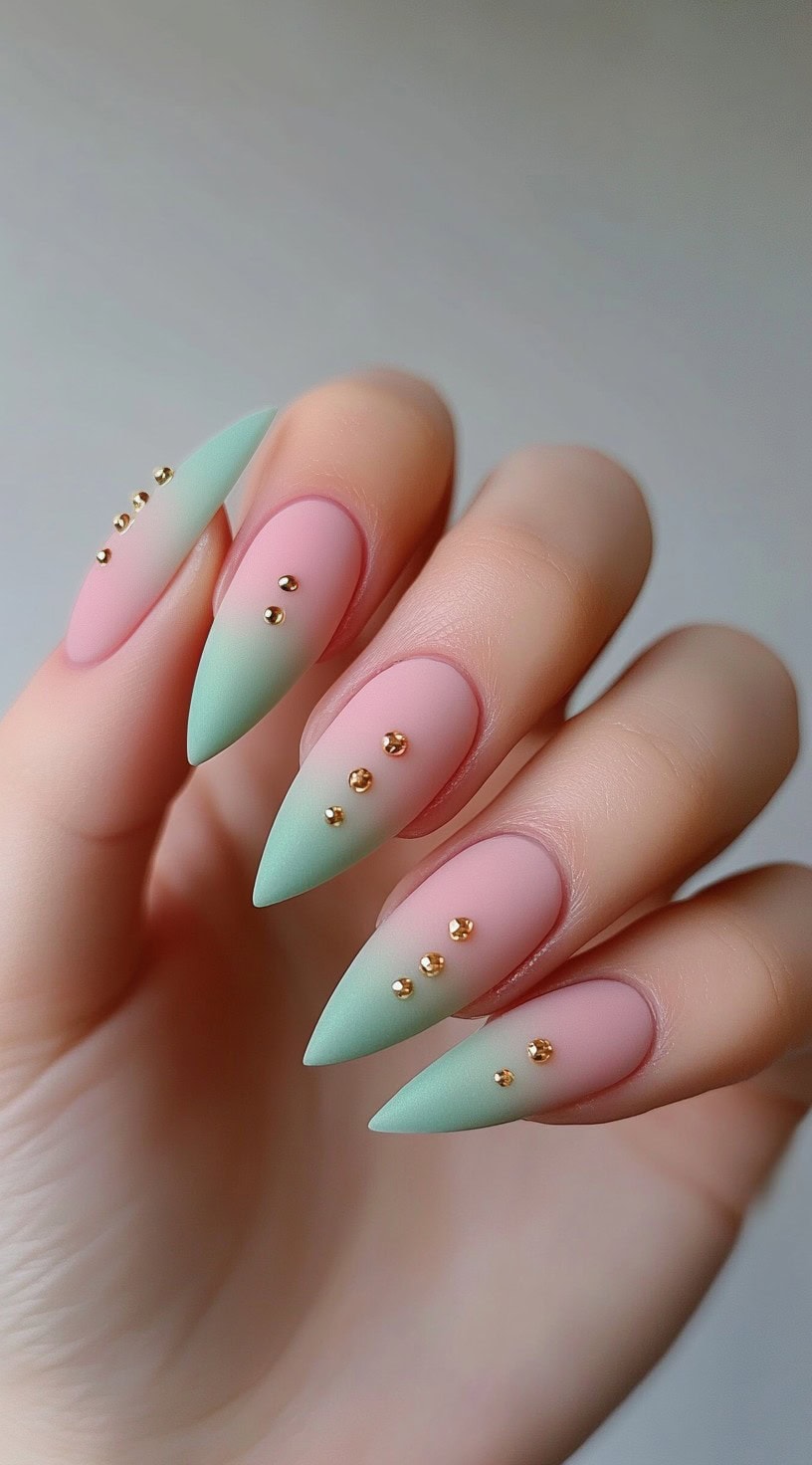 A hand with matte pink and green ombre stiletto nails decorated with tiny gold studs.