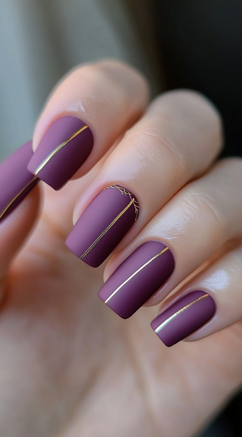 Matte plum square nails decorated with thin gold vertical striping lines, one nail accented with crosshatch gold detail.