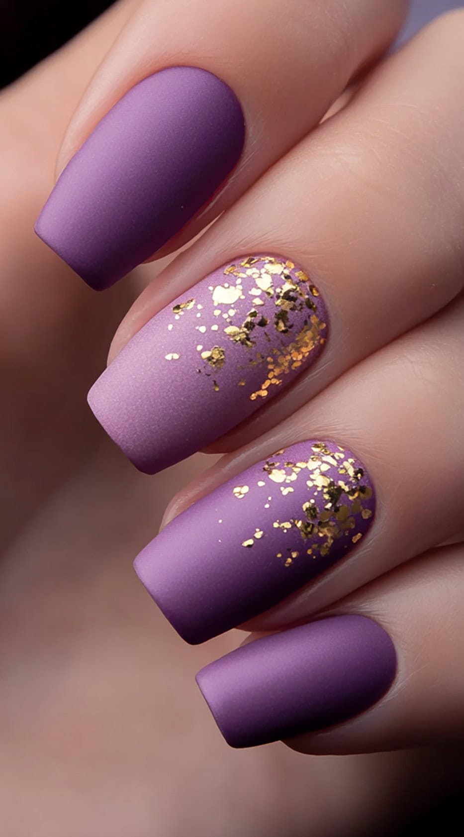 A hand with square-shaped nails in matte purple ombré, featuring gold foil accents on two fingers.