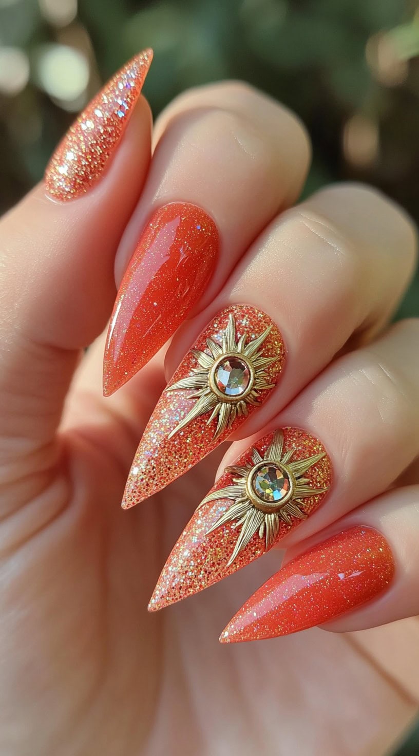 Hand with long stiletto nails painted in orange glitter and adorned with 3D golden sun charms with crystals.