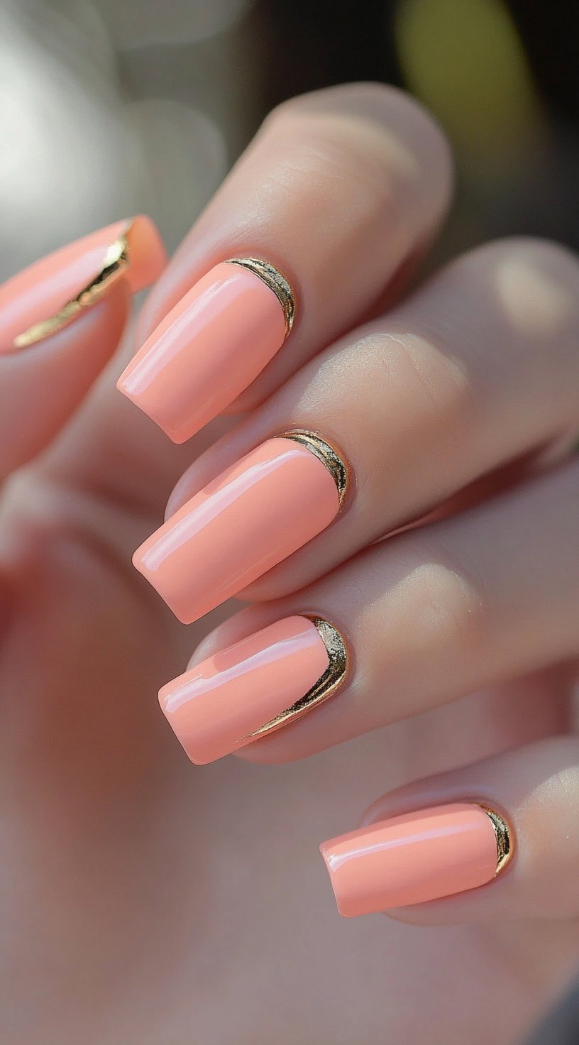 A close-up of square-shaped nails painted in glossy peach nude with metallic gold half-moon detailing near the cuticles.