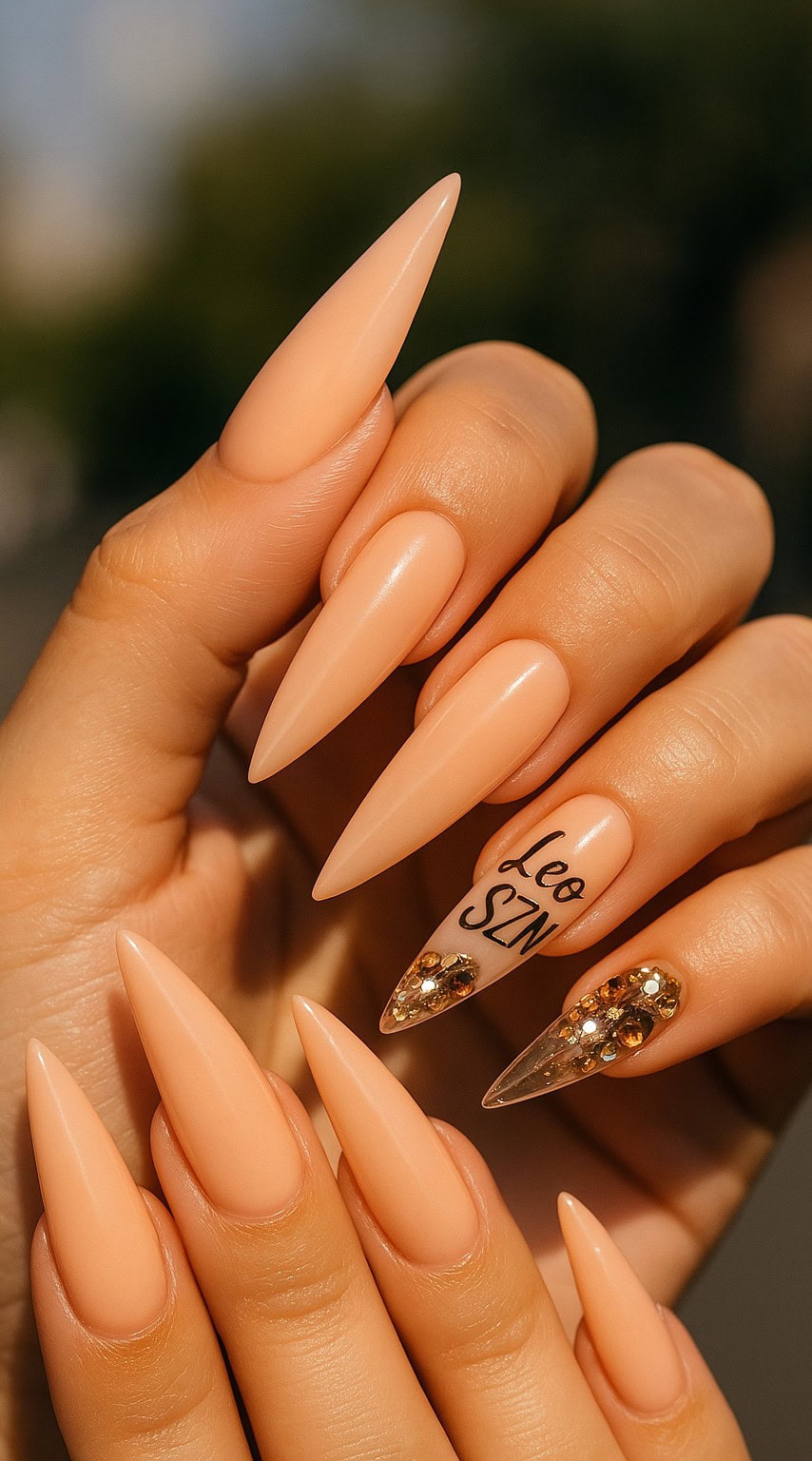 Long stiletto nails in peach tone, with 'Leo SZN' text and gold rhinestone accents on clear nails.