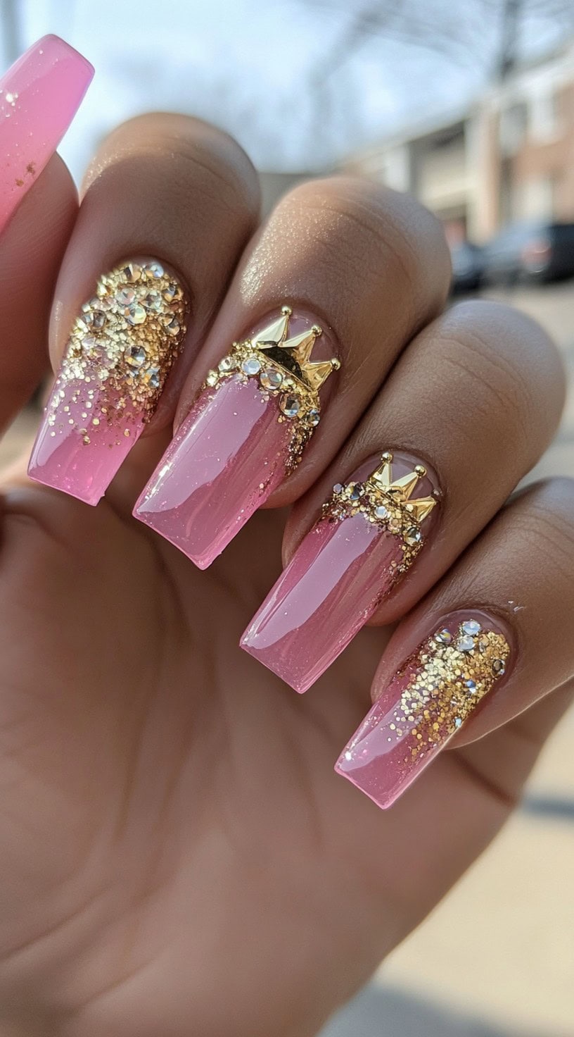 Square nails with pink polish, gold glitter accents, and golden crown charms surrounded by rhinestones.
