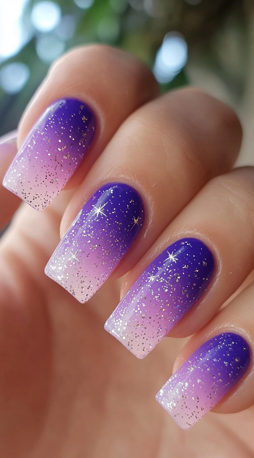 Square-shaped nails with a gradient of lavender to deep blue and scattered silver glitter.