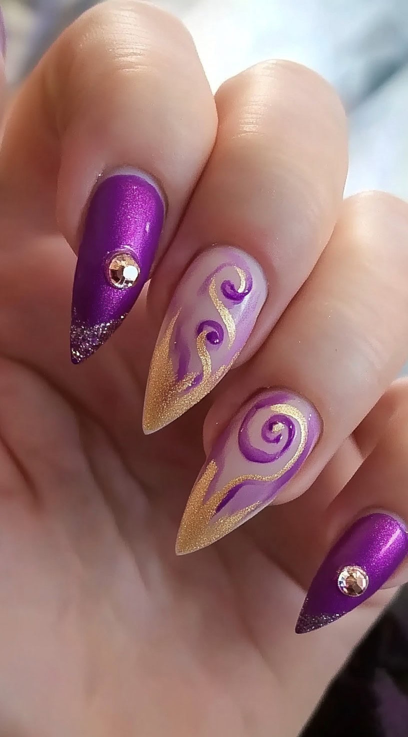 Stiletto nails with a mix of matte and glossy purple polish, gold glitter swirls, and rhinestone embellishments on the outer nails.