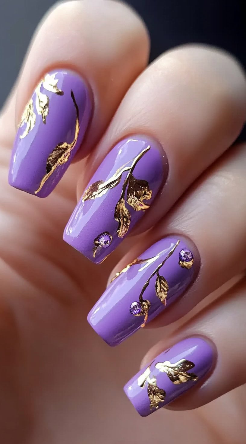 Square nails in a lilac-purple tone with detailed gold foil branches and small crystal accents.