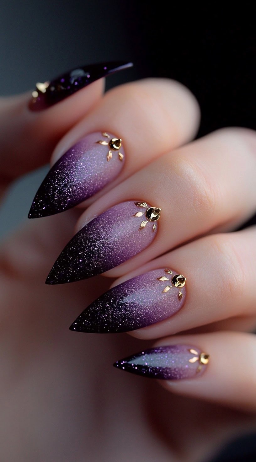 Almond-shaped nails in a gradient of purple glitter with small gold sunburst studs near the cuticles.