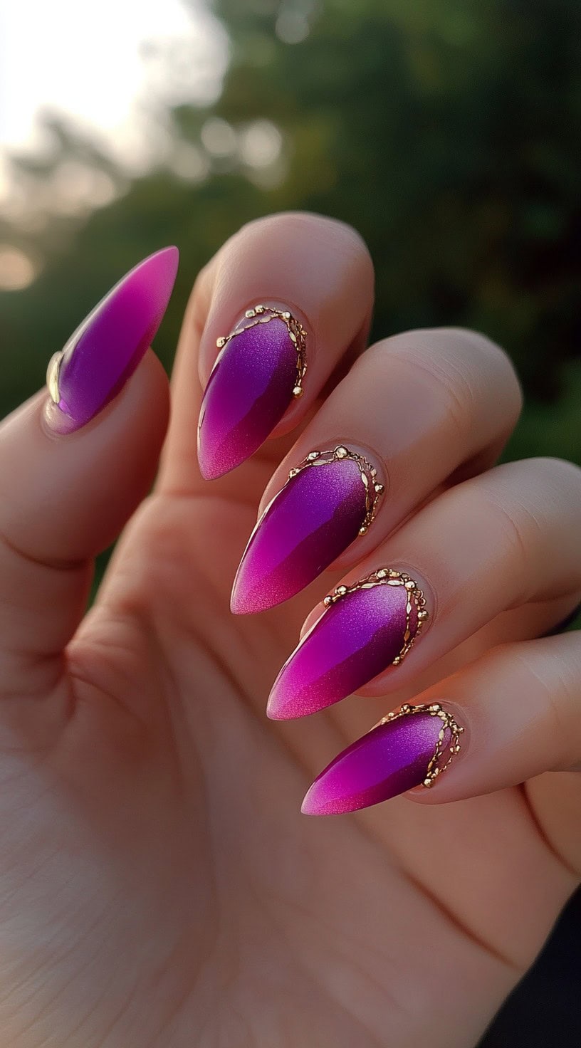 Stiletto nails with a vibrant pink-to-purple ombre and ornate gold detailing around the base.
