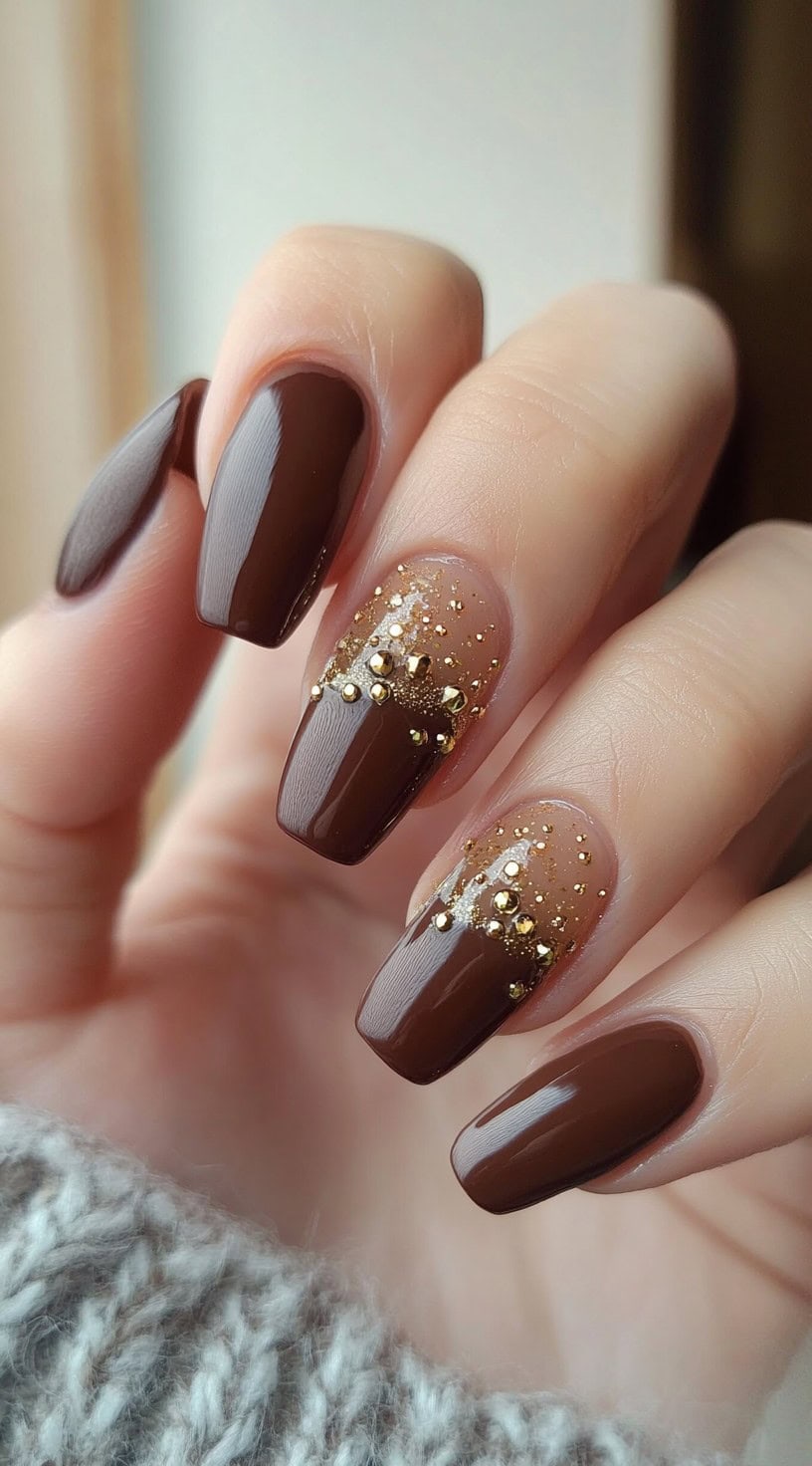 Square-shaped nails painted in a deep brown with gold glitter accents and studded embellishments on two feature nails.