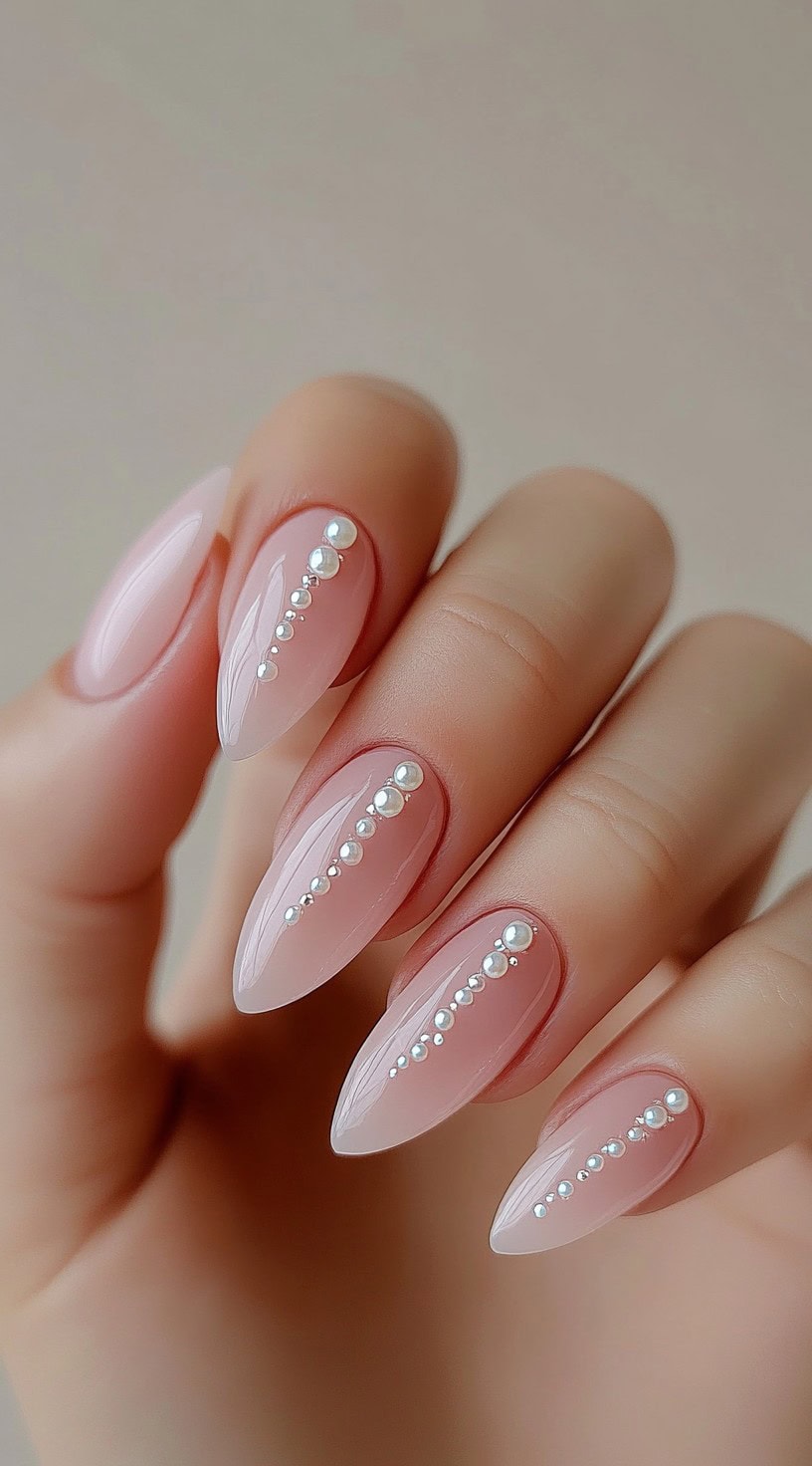 A close-up of almond-shaped nails painted in a sheer pink color, adorned with a vertical line of small pearls down the center of each nail.