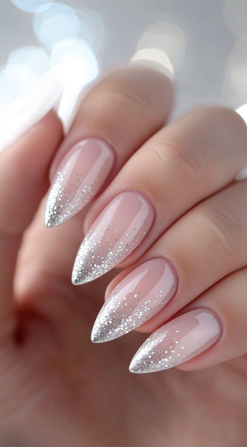 A set of long, almond-shaped nails with a soft pink base transitioning into silver glitter ombre tips.