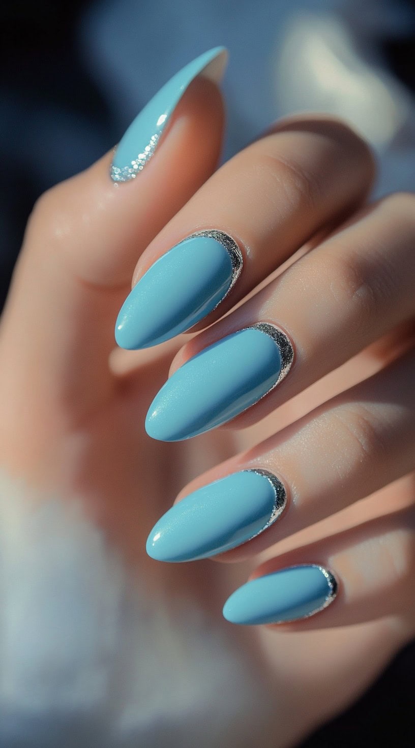 A hand with almond-shaped nails painted in sky blue and outlined with fine silver glitter along the cuticle edge.