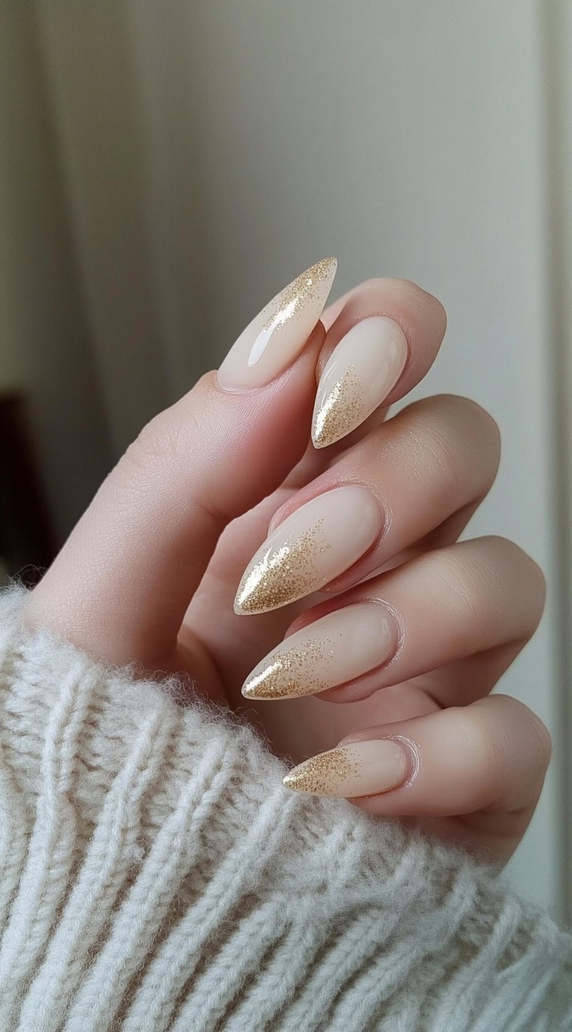 A close-up of long almond-shaped nails with a sheer nude base and gold glitter fading from the tips downward.
