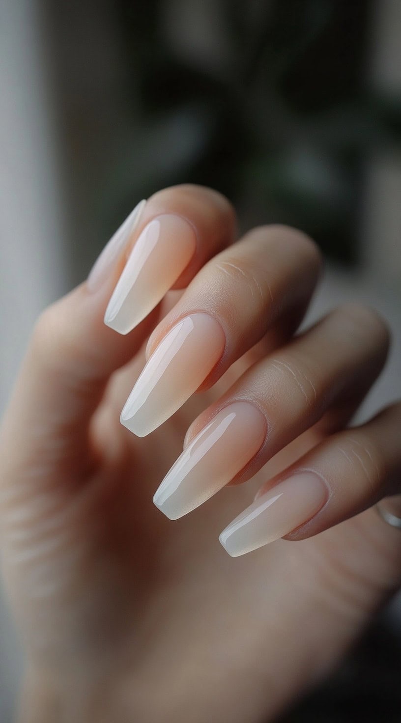 A hand with long, coffin-shaped nails featuring a soft nude-to-white ombre effect with a glossy finish.