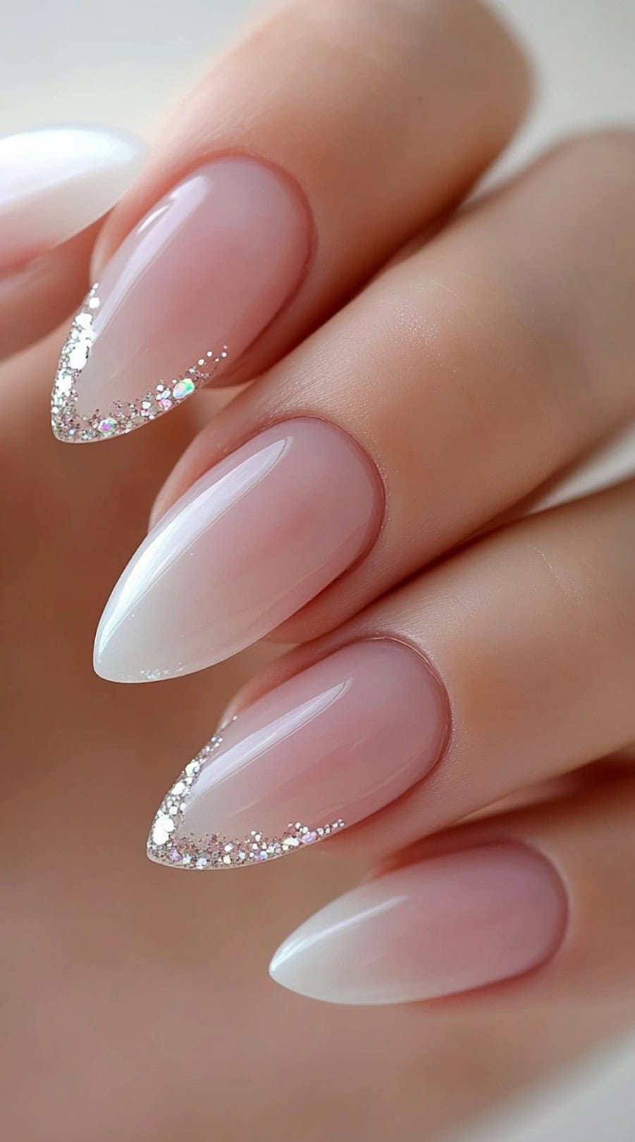 A hand with almond-shaped nails in a sheer pink ombré design, topped with fine silver glitter along the tips.