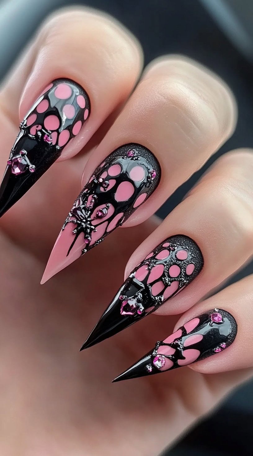 A hand with long stiletto nails featuring a webbed black and pink design with spider and gem embellishments.