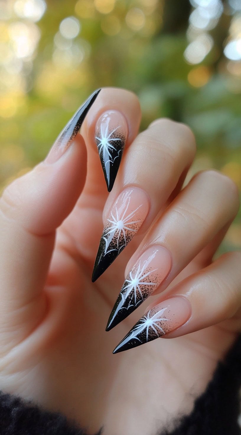 Stiletto nails with a black-to-clear ombre base and hand-drawn white spiderweb starburst designs.