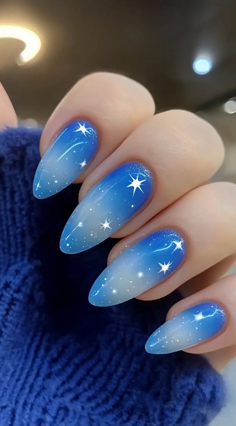 Oval-shaped nails featuring a celestial blue aura with scattered white star details over a sheer ombré base.