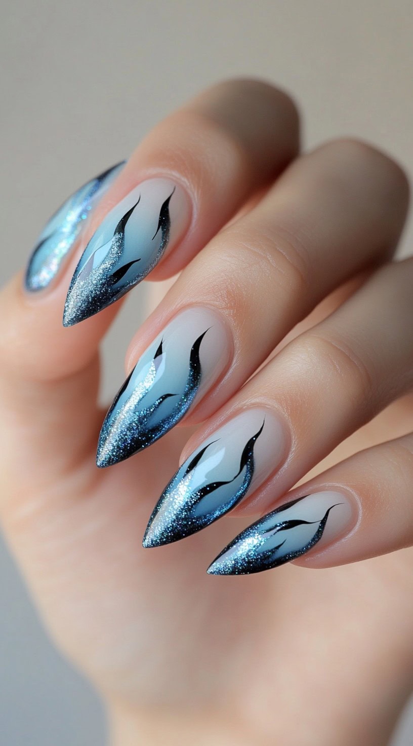 Long stiletto nails with a gradient blue glitter ombre design accented by black flame details.