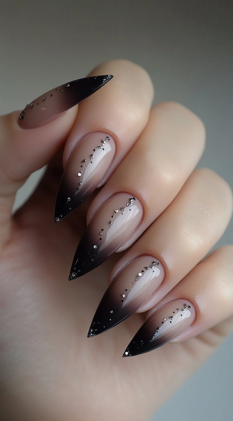 Long stiletto nails with a nude-to-black ombre fade and delicate rhinestones flowing diagonally across each nail.