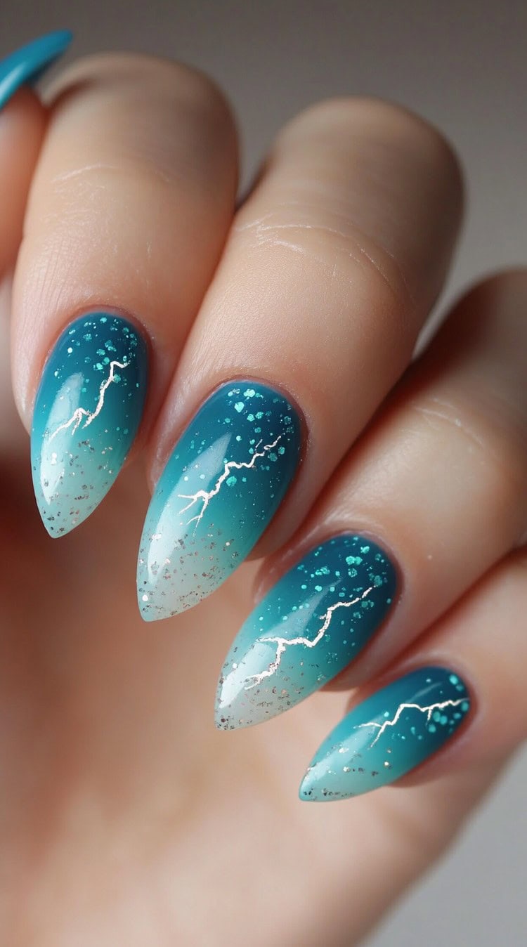 Almond-shaped nails with a teal to pale blue ombre, scattered glitter, and silver lightning-like accents.