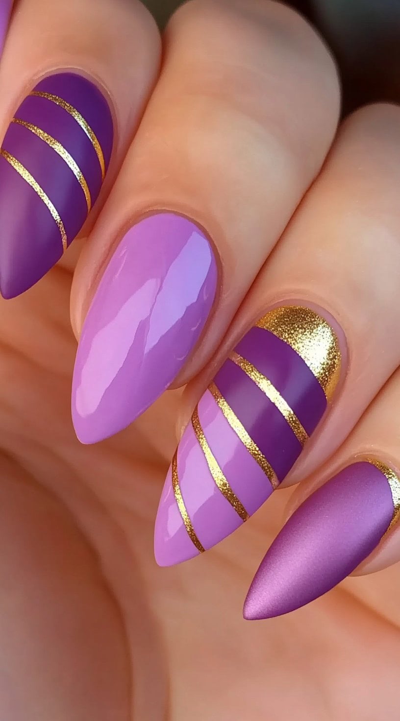 Long almond nails painted in alternating glossy and matte purples with bold diagonal gold stripe accents.
