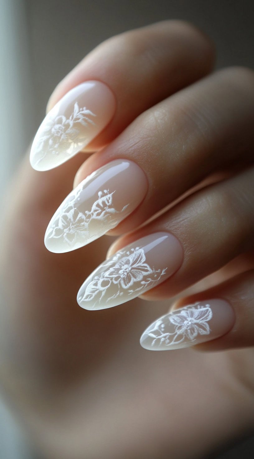 A set of almond-shaped nails with a soft nude base, featuring intricate white floral lace designs extending from the tips.